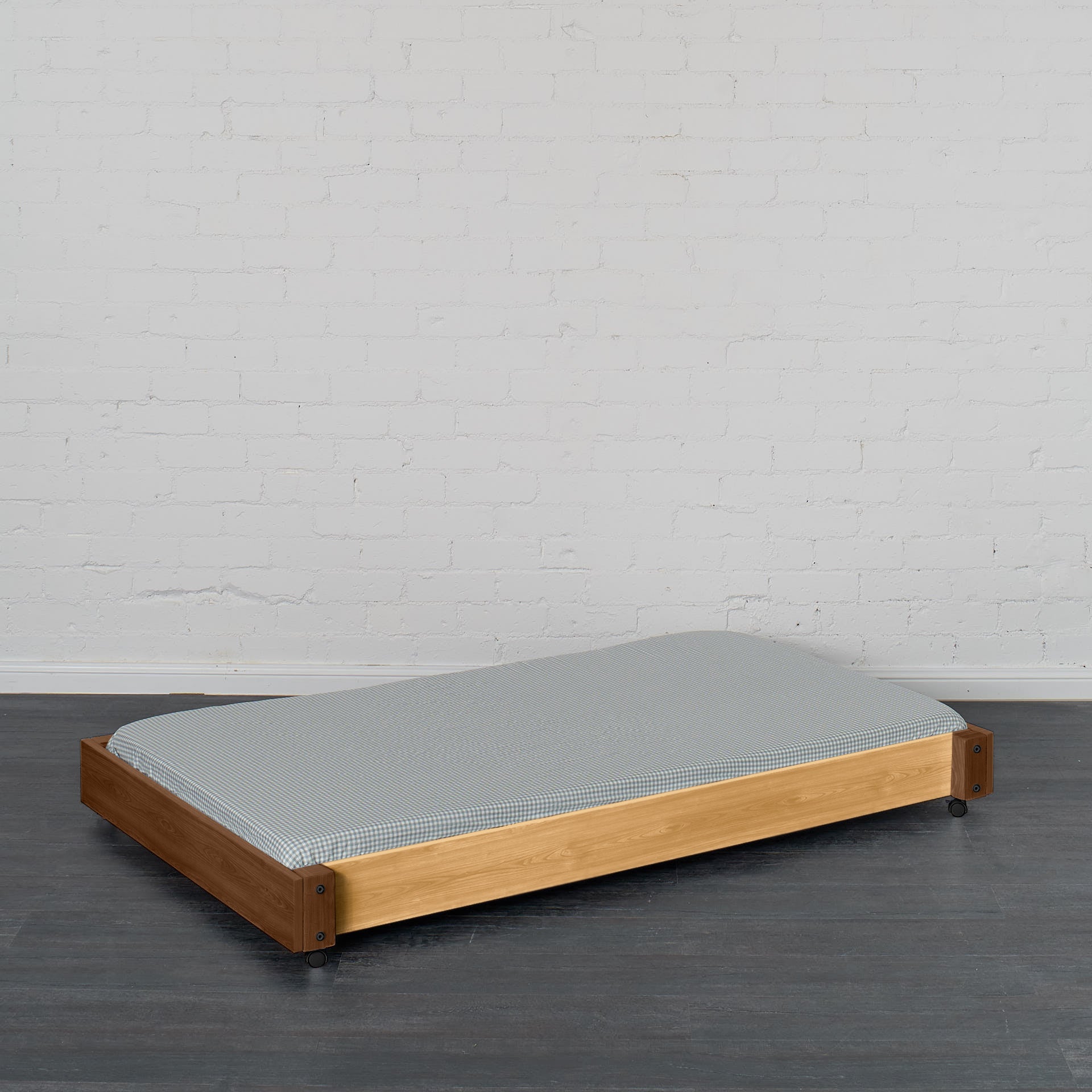 Low wooden trundle bed frame in natural elm with gray mattress, designed to slide under a bunk or loft bed.