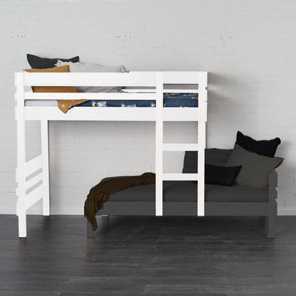 White mid-line loft bed with safety rails and ladder over a dark sofa bed against a white wall.