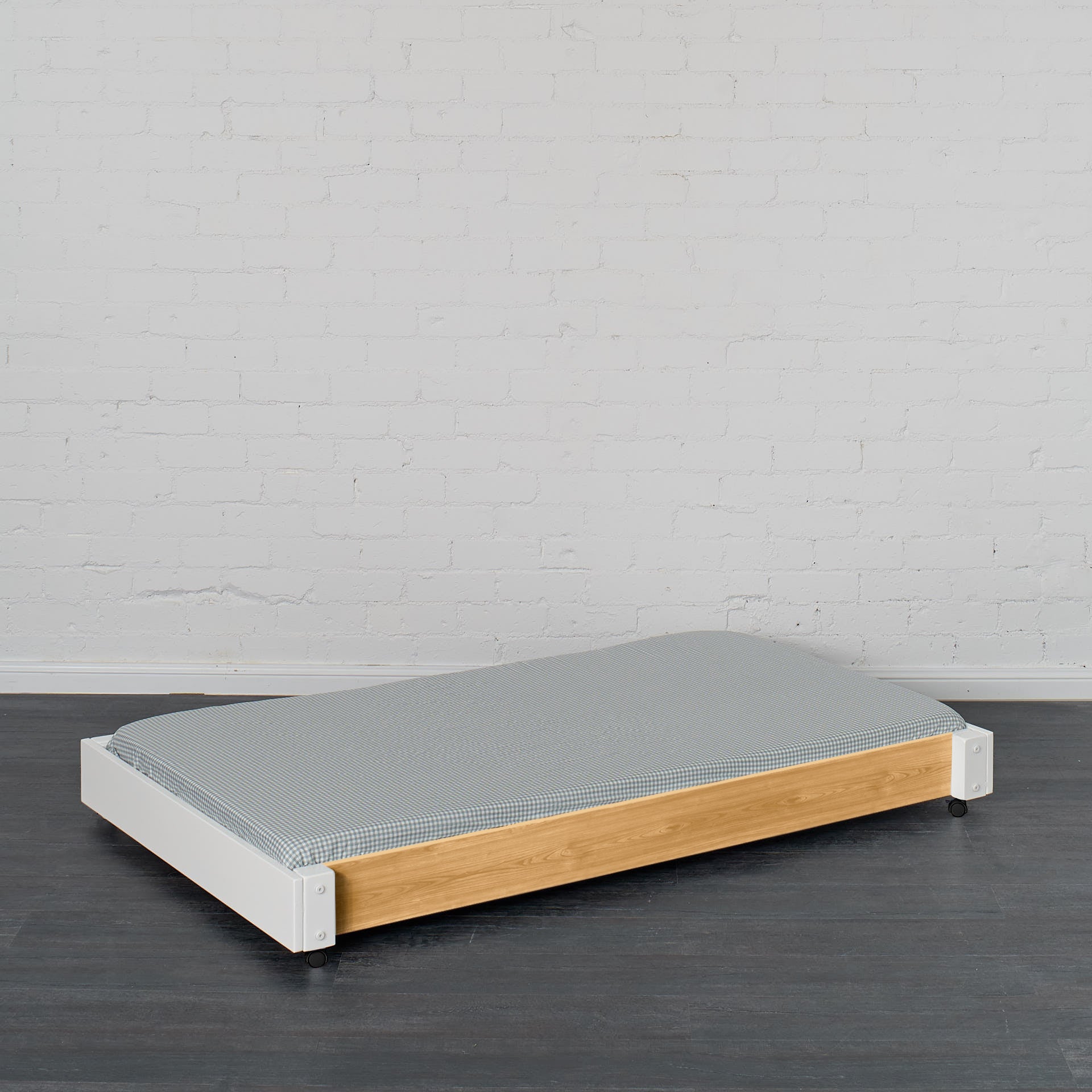 Low wooden trundle bed with white frame and grey mattress on dark floor.