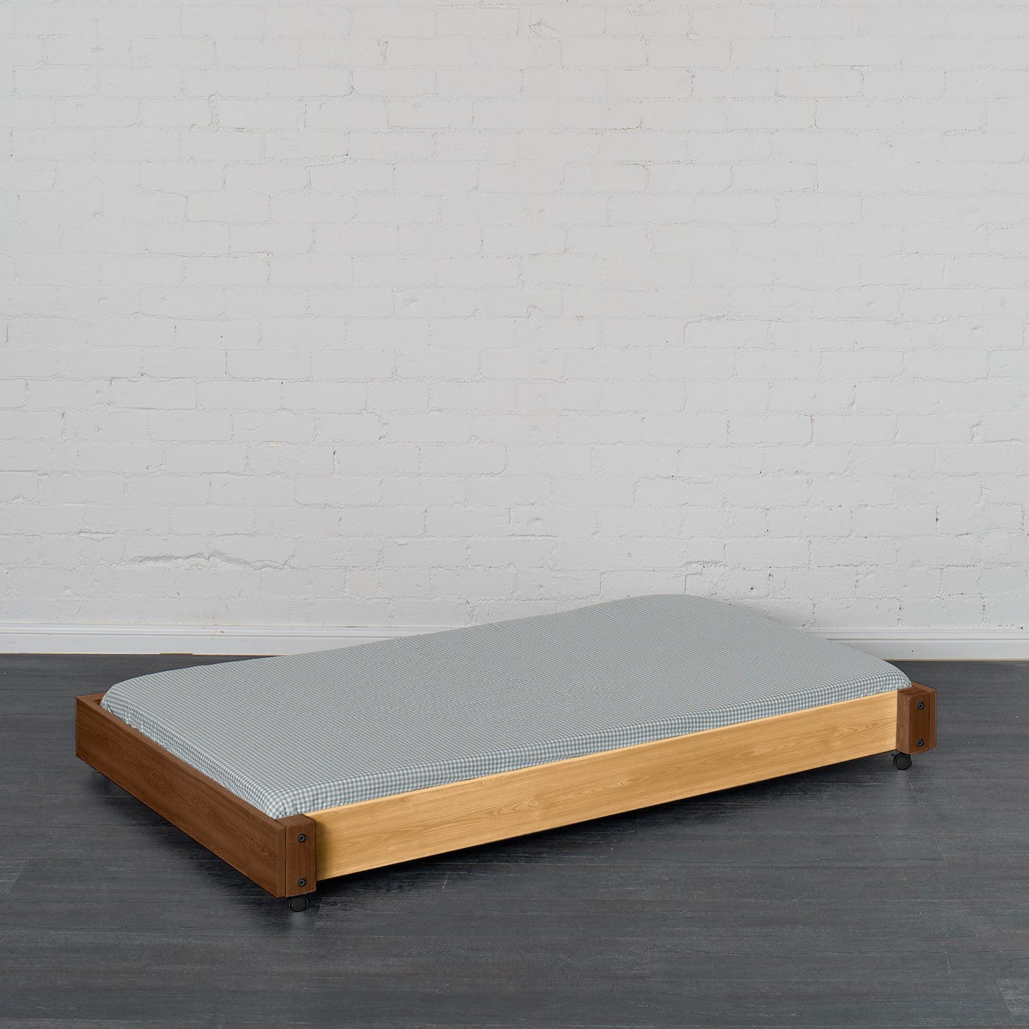 Low wooden trundle bed frame in natural elm with gray mattress, designed to slide under a bunk or loft bed.