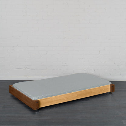 Low wooden trundle bed frame in natural elm with gray mattress, designed to slide under a bunk or loft bed.